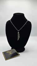 Silver necklace with feather pendant on a black bust, next to a branded card on a white background