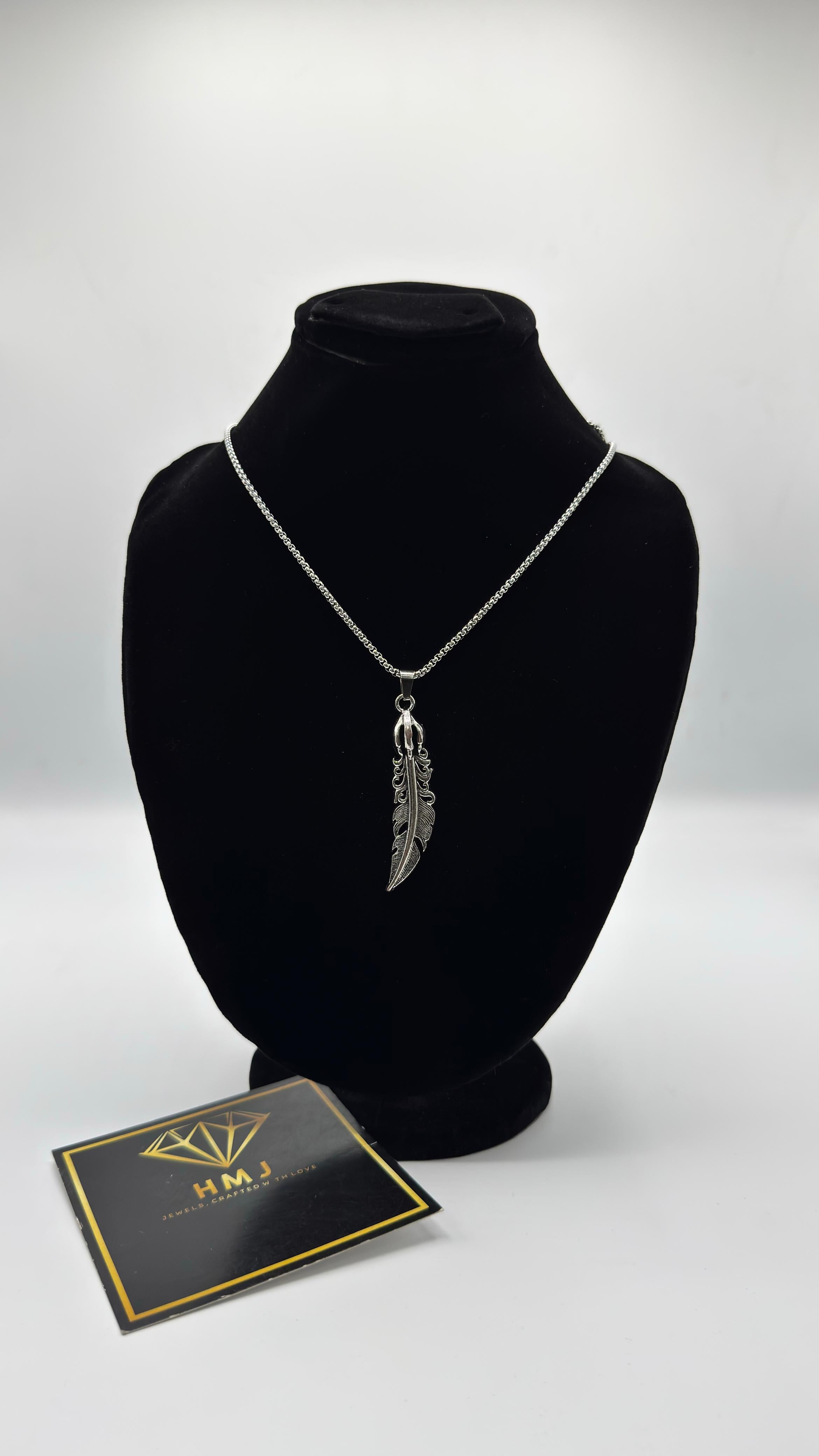 Silver necklace with feather pendant on a black bust, next to a branded card on a white background
